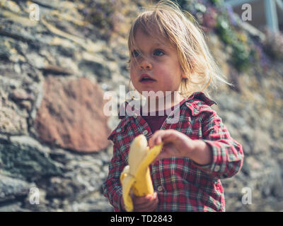 Un peu de tout-petit est manger une banane à l'extérieur par un mur en pierre Banque D'Images