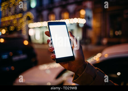 Woman's hand holding mobile phone avec écran blanc sur fond de route avec les voitures et nuit au chaud lit city, close-up. Banque D'Images