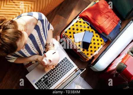 Vue supérieure de l'élégante femme en pantalon blanc et chemisier rayé dans la maison moderne en journée ensoleillée holding piggy bank près de l'open travel suitcase pla Banque D'Images