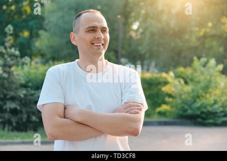 Outdoor portrait of smiling sporty homme de 40 ans. Beau mâle positive avec les bras croisés en t-shirt blanc, le fond est le coucher du soleil d'été je Banque D'Images