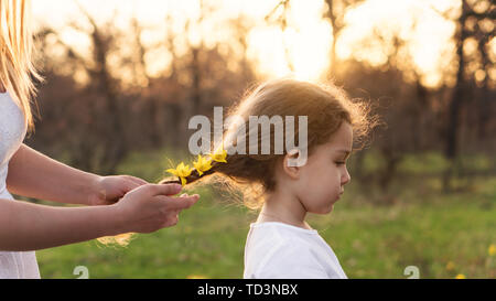 Prendre soin des mains maternelle braid les cheveux de petite fille. Maman décore ses cheveux en tressage jaune fleurs forêt vivent en fibre amorce. Professionnels de la maternité. Enfant Banque D'Images