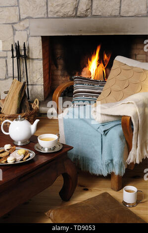 Théière et biscuits sur une table avec une chaise en face d'une cheminée avec du feu allumé seulement droits britannique et irlandais Banque D'Images