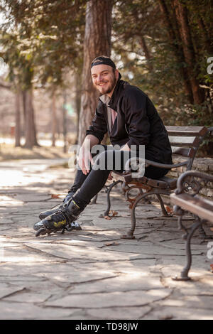 Un jeune homme souriant, assis sur un banc dans un parc, le port de patins. Banque D'Images