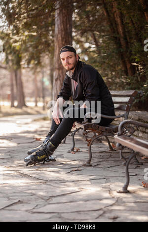 Un jeune homme avec sérieux, assis sur un banc dans le parc, le port de patins. Banque D'Images