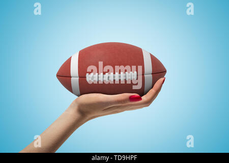 Gros plan du côté de woman's hand holding dirigé vers le haut et big brown ball pour le football américain sur fond dégradé bleu clair. Banque D'Images