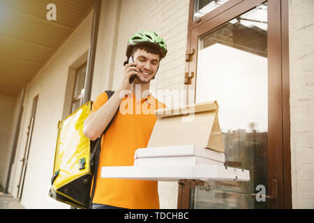 Jeune homme en chemise jaune livraison pizza à l'aide de gadgets pour suivre la commande à la rue. L'utilisation de messagerie en ligne l'app pour recevoir le paiement et le suivi de l'adresse d'expédition. Les technologies modernes. Banque D'Images