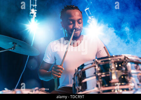 Jeune musicien de jazz afro-américains ou le batteur jouant des percussions sur fond bleu lumineux studio dans la fumée autour de lui. Concept de la musique, passe-temps, inspirness. Portrait coloré de l'artiste. attrayant joyeux Banque D'Images