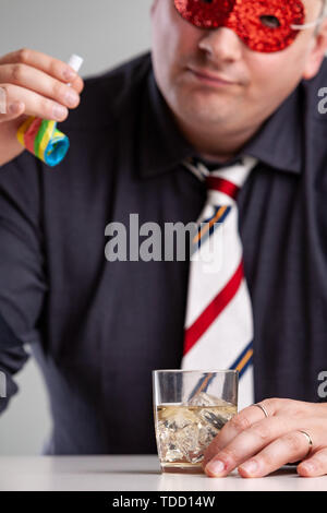 Un homme portant un masque de fête colorés assis boire de l'alcool lors d'une fête à la recherche d'un ventilateur ou d'un parti à corne avec un expression dans un close up portrait Banque D'Images