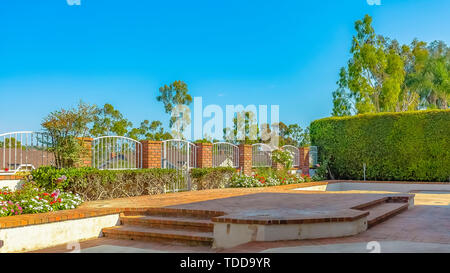 Patio de brique Panorama avec une plate-forme surélevée à l'avant de l'arche portail blanc. Hauteur d'une haie, de plantes à fleurs et arbres luxuriants entourent la région. Banque D'Images