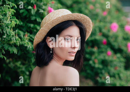 Piscine close up portrait of young beautiful happy smiling girl wearing straw hat élégant. Model posing près de rosiers. @Summer mode concept. Banque D'Images