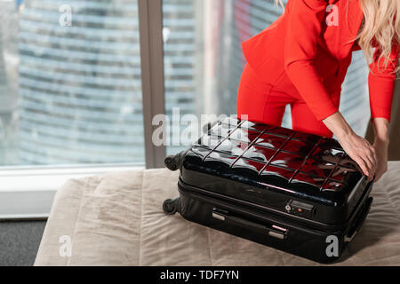 Close-up dépaquète valise jeune blond businesswoman arrive dans une chambre d'hôtel avec valise noire. Femme en costume d'affaires de corail rouge. Banque D'Images