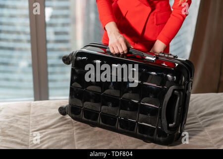 Close-up dépaquète valise jeune blond businesswoman arrive dans une chambre d'hôtel avec valise noire. Femme en costume d'affaires de corail rouge. Banque D'Images