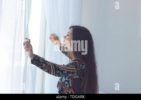 Belle jeune femme poussant les rideaux translucides sur la fenêtre. Banque D'Images