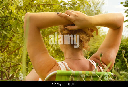 Vue arrière, jeune femme assise sur une chaise de repos dans le jardin et en maintenant ses mains derrière sa tête, belle femme chef profil par rapport à la zone de Banque D'Images