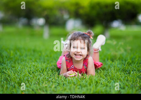 Happy Smiling Little Girl Laying On Grass In Park avec robe rouge. Banque D'Images