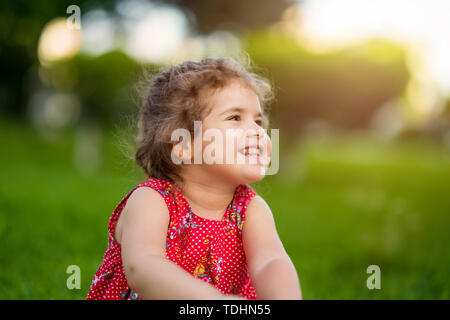 Happy Smiling Little Girl Sitting on Grass In Park avec robe rouge. Banque D'Images