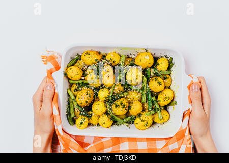 Woman's hands holding de cuisson avec un plat fait maison fraîchement préparé avec des pommes de terre nouvelles au four haricots verts sur fond blanc, vue d'en haut. Banque D'Images