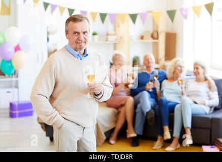 Portrait of content beau vieil homme avec barbe portant chandail occasionnels de flûte à champagne d'anniversaire alors que ses friends chatting Banque D'Images