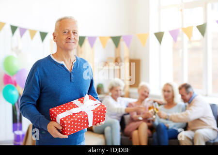 Portrait de l'homme anniversaire beau contenu dans pull bleu debout dans la salle de séjour décorée pour anniversaire et holding gift box pendant que ses amis Banque D'Images
