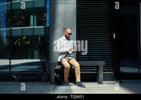 Jeune homme barbu concentrées dans des lunettes de soleil assis sur un banc et la vérification des fichiers en ligne sur tablette Banque D'Images