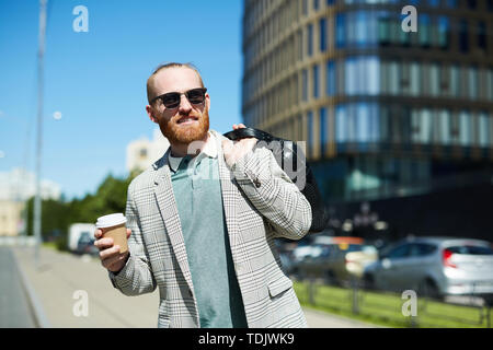 Smiling handsome red-bearded man in sunglasses holding karate cup et du portefeuille sur le dos et à distance dans la cabine en attendant on st Banque D'Images