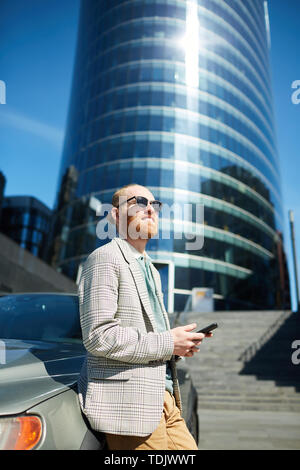 Une jeune hipster positive entrepreneur barbu à lunettes s'appuyant sur en voiture du quartier financier et des sms message sur téléphone portable Banque D'Images