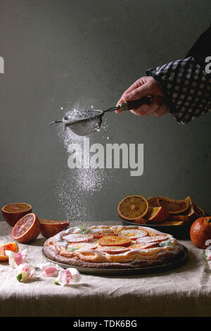 La main du jeune homme du sucre en poudre d'arrosage des Cheesecake avec orange sanguine de Sicile, décoré par des fleurs comestibles, les feuilles de menthe servi sur la ligne grise Banque D'Images
