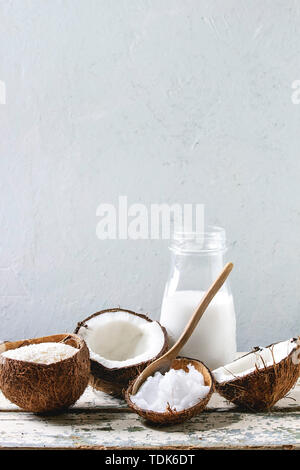 Gamme de produits de la noix de coco lait en bouteille de verre, et l'huile, en coquille, frais de flocons de noix de coco cassées sur la vieille table en bois avec mur gris à l'arrière-plan. Guérir Banque D'Images