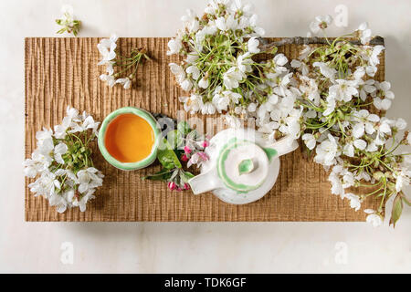 Fait main artisanat théière et tasse en céramique avec du thé chaud décoré par des branches de cerisier en fleurs au printemps sur le plateau de service en bois sur fond de marbre blanc. Fl Banque D'Images