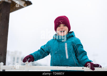 Happy Baby smiling. Le garçon joue dans l'hiver dans l'aire de jeux. l'enfant est vêtu d'un chapeau rouge, bleu veste et mitaines Banque D'Images