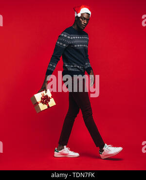 Young African man wearing santa hat sautant avec boîte-cadeau sur fond rouge. Beau black avec un cadeau de Noël sur fond rouge. Banque D'Images