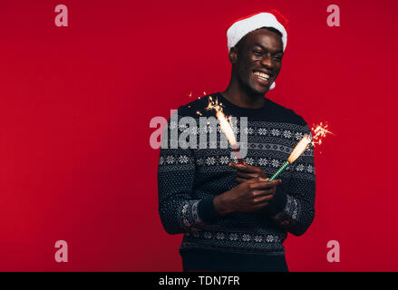 Cheerful young African man in santa hat fête avec baguettes. Jeune homme jouissant avec Fireworks sur fond rouge Banque D'Images