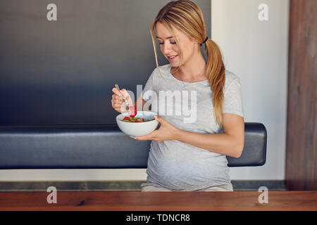 Jeune femme enceinte bénéficiant d'une bonne salade de fruits frais en souriant joyeusement à la caméra comme elle tient le bol dans sa main en prenant une bouchée assis à Banque D'Images