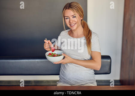 Jeune femme enceinte bénéficiant d'une bonne salade de fruits frais en souriant joyeusement à la caméra comme elle tient le bol dans sa main en prenant une bouchée assis à Banque D'Images