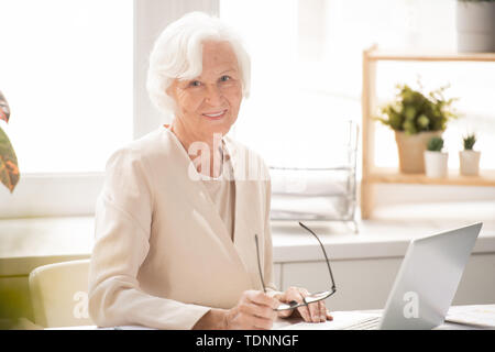 De smiling businesswoman in costume élégant blanc vous regarde en position assise par bureau en face de laptop in office Banque D'Images