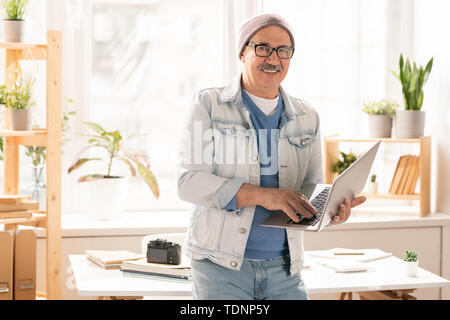 Homme mature en souriant veste en jean bleu jeans et pull à l'article 24 et par la saisie sur clavier d'ordinateur portable in office Banque D'Images