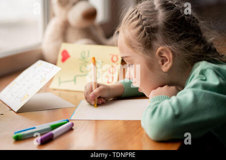 Concentré grave fille aux cheveux tressés en s'appuyant sur une table en bois et en carte postale pour papa sur fête des pères Banque D'Images