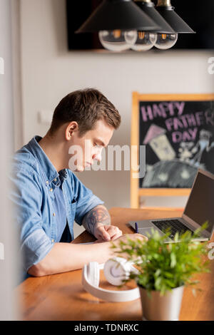 Jeune homme concentré freelancer en chemise en jean assis à table avec ordinateur portable et les tâches d'écriture vers le bas, heureuse fête des pères dessin sur tableau noir à l'arrière Banque D'Images