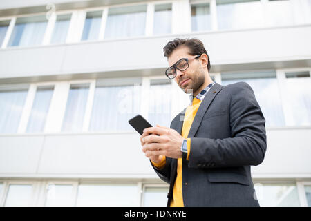 Jeune homme sérieux ou avocat dans smart casual textos ou défilement dans smartphone dans l'environnement urbain Banque D'Images