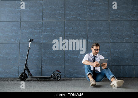 Jeune homme détendu dans smart casualwear assis au mur du bâtiment et le défilement dans digital tablet Banque D'Images