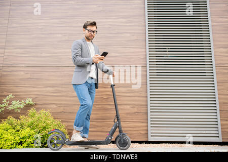 Les jeunes de l'employé mobile sérieux dans une tenue de garder une jambe sur un scooter tout en envoyant dans le smartphone dans l'environnement urbain Banque D'Images