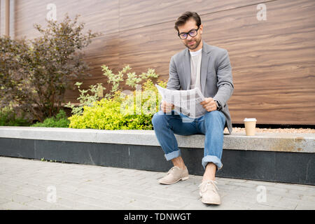 Jeune homme urbain en jeans et veste assis dans l'environnement urbain et lisant le journal avec les dernières nouvelles Banque D'Images