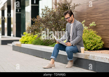 Jeune entrepreneur de smart casual assis dans l'environnement urbain et à l'intermédiaire de journal avec des nouvelles fraîches Banque D'Images