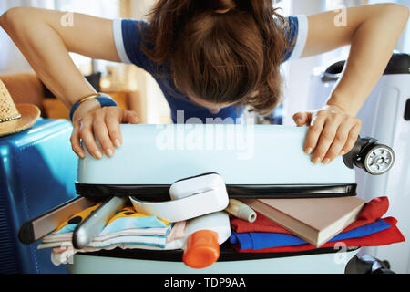 La femme en bleu voyageur élégant T-shirt à la maison moderne en journée ensoleillée essayant de fermer plus de paniers-valise. Banque D'Images