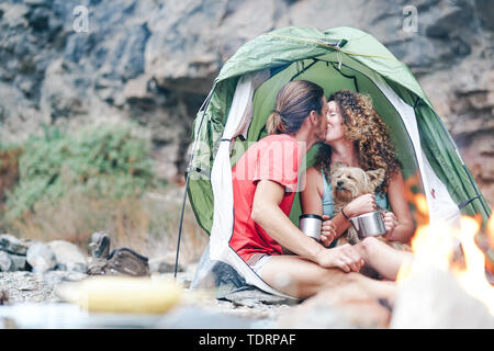 Couple voyage en camping dans les montagnes de roche après une journée de trekking - Heureux les personnes qui boivent du thé chaud à l'intérieur de tente avec son chien à côté de feu Banque D'Images