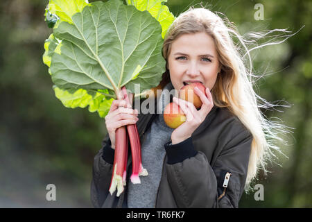 Rhubarbe (Rheum rhabarbarum), jeune femme blonde avec de la rhubarbe a mordre avec plaisir dans une pomme, Allemagne Banque D'Images