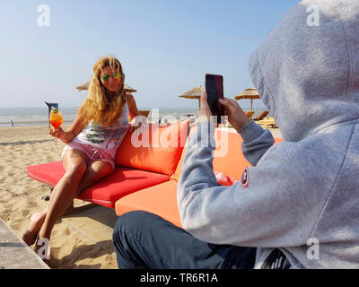 Sweat homme avec prise de photos d'une femme blonde avec verre à cocktail sur la plage de sable, Pays-Bas Banque D'Images