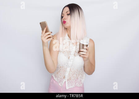 Image de jolie belle jeune femme blonde en chemise blanche posant sur fond blanc à boire du café et à l'aide de son shartphone Banque D'Images