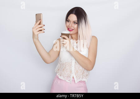 Image de jolie belle jeune femme blonde en chemise blanche posant sur fond blanc à boire du café et à l'aide de son shartphone Banque D'Images