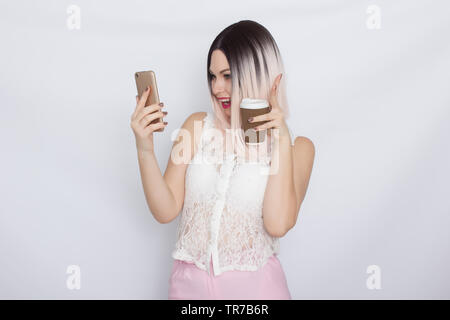 Image de jolie belle jeune femme blonde en chemise blanche posant sur fond blanc à boire du café et à l'aide de son shartphone Banque D'Images